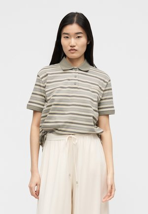 Tânără cu păr lung și închis la culoare, purtând un tricou polo cu mâneci scurte, cu dungi, și pantaloni largi, de culoare bej deschis, pe un fundal simplu.