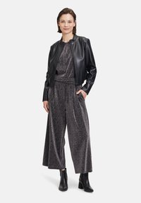 Schwarze Leder-Bomberjacke über einem glänzenden, strukturierten, grauen Jumpsuit mit weitem Bein. Das Model trägt schwarze Knöchelstiefel und steht mit den Händen in den Taschen.