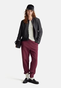 Giacca nera corta, maglietta bianca a costine, pantaloni sportivi bordeaux, mocassini neri, calzini bianchi e un cappellino nero con logo, mentre tiene una borsa nera.