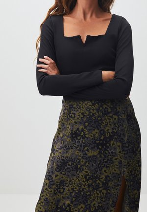 Femme les bras croisés portant un haut noir à manches longues avec une encolure carrée et une jupe à motifs avec une fente latérale.