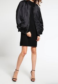 Svart satinbombarjacka med ribbade cuffar, bärs över en svart klänning. Svart högklackade sandaler med remmar fulländar outfiten.