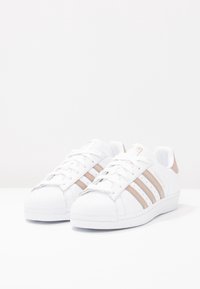 Sneakers bianchi con punta textured, tre strisce rosé in oro e tomaia in pelle. Suola in gomma e colletto imbottito per il comfort.