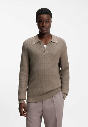 Mand iført beige, struktureret striktrøje med krave og to knapper, kombineret med lyse taupebukser, stående mod en hvid baggrund.