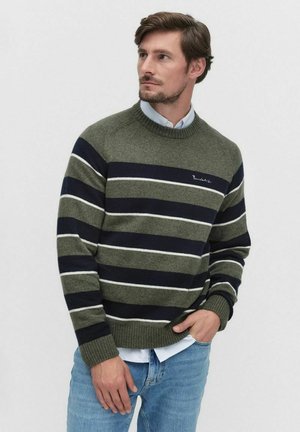 Man in een olijfgroene en marineblauwe gestreepte trui over een lichtblauw shirt, met een spijkerbroek, die naar de zijkant kijkt met zijn hand in zijn zak.