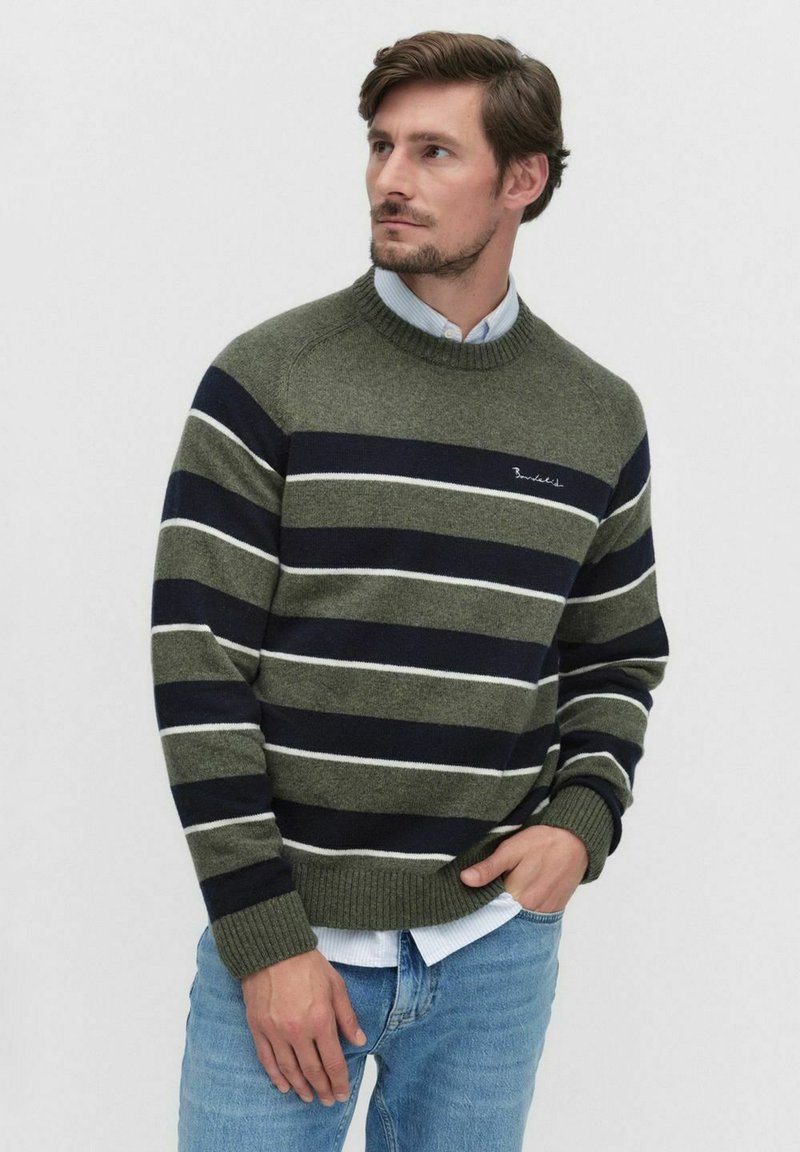 Man in een olijfgroene en marineblauwe gestreepte trui over een lichtblauw shirt, met een spijkerbroek, die naar de zijkant kijkt met zijn hand in zijn zak.