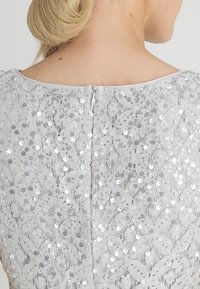 Robe en dentelle à sequins argentés avec un motif floral détaillé, texture douce, encolure ronde et fermeture à zip au dos. Couleur : gris clair.