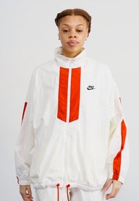 Fehér széldzseki piros kiegészítőkkel és csíkokkal, magas gallérral, állítható derékpánttal, és fekete Nike logóval a mellkason.