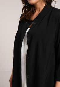 Veste noire de style kimono en tissu lisse, dotée d'un col mandarin et de boutons ronds, portée par-dessus un top blanc.
