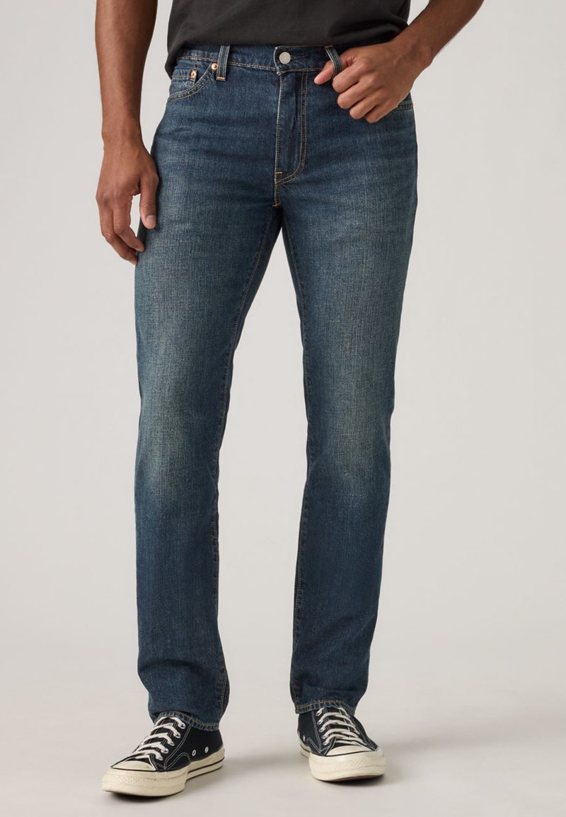 Homme portant un jean slim bleu foncé et des baskets basses noires, debout devant un fond clair uni.