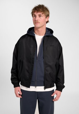 Mężczyzna z jasnobrązowymi włosami i wąsami, ubrany w czarną kurtkę zapinaną na zamek błyskawiczny, na granatowej bluzie z kapturem i białej koszulce, stojący na białym tle.