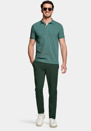Uomo che indossa una polo verde scuro, pantaloni verde scuro, sneakers bianche e occhiali da sole, sorridente con una mano in tasca, in piedi su sfondo bianco.