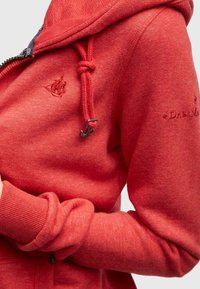 Rød fleece hættejakke med lynlås, snoretræk i hætten og broderet logo. Har ribbede manchetter og en blød tekstur.