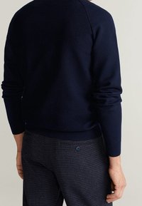 Pull texturé bleu marine avec col rond et poignets côtelés, associé à un pantalon à carreaux foncés avec une poche arrière boutonnée.