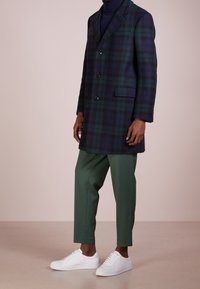 Cappotto doppiopetto a quadri in verde scuro e blu navy, tessuto in lana, con rever a incavo, tre bottoni, tasche laterali; abbinato a pantaloni verdi e sneakers bianche.