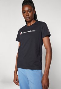 Černé bavlněné tričko s krátkým rukávem, s bílým nápisem "Champion" napsaným napříč hrudníkem a logem na levém rukávu.