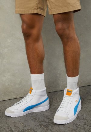 Baskets montantes blanches avec une bande latérale bleue, des accents de logo orange et des semelles texturées. Portées avec des chaussettes blanches et un short kaki.