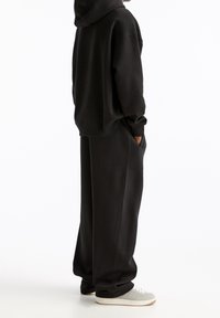 Zwarte oversized hoodie met een voorzak en bijpassende wijde sweatpants. Het materiaal lijkt zacht met een gladde textuur. Grijze sneakers maken de outfit compleet.
