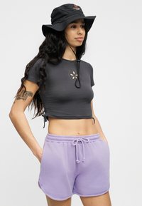Νεαρή γυναίκα με μακριά σκούρα μαλλιά, φοράει μαύρο καπέλο τύπου μπάκετ, σκουρό γκρι κολλητό cropped t-shirt με λουλουδάτο άνοιγμα και ανοιχτό μωβ σορτς με κορδόνια στη μέση.