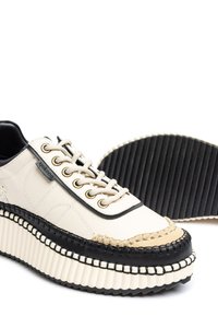 Crème en zwarte platform sneaker met dikke ribbelzool, vetersluiting aan de voorkant, zichtbare stiksels en een merklabel aan de zijkant.