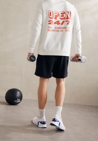 Sudadera blanca con texto rojo "ABIERTO 24/7", pantalones cortos negros, zapatillas blancas con detalles azules y pesas en las manos. Una bola medicinal negra cerca.