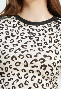 Tričko s leopardím potiskem vyrobené z lehkého materiálu. Černobéžový vzor s černým žebrovaným výstřihem a logem Adidas ve středu.