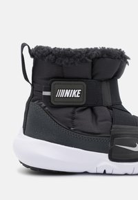 Svart vadderad hög top-sneaker med pälskrage, kardborreband och vit gummisula; har Nike-logotyp och strukturerade detaljer.