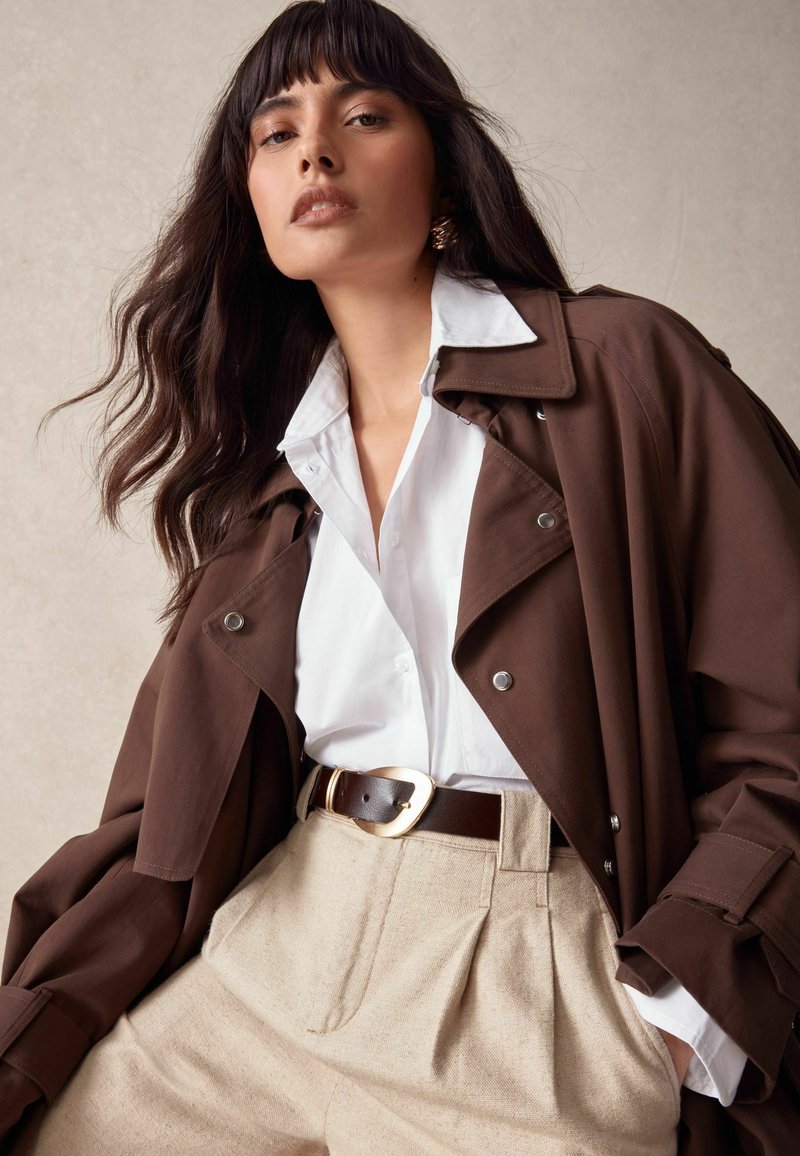 Brun trenchcoat över en vit skjorta med knappar, beige veckade byxor och ett brunt bälte med ett guldspänne. Mjuk tyg, avslappnad passform.