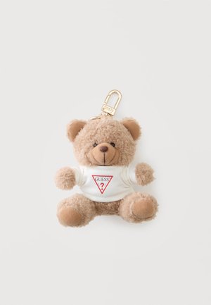 Llavero pequeño de osito de peluche beige que lleva una camiseta blanca con un logo rojo de Guess y un cierre metálico dorado.