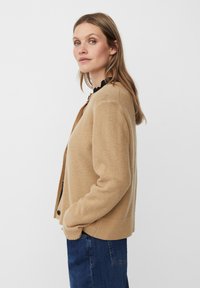 Beige gestrickte Strickjacke mit gerippten Bündchen und Saum, mit rundem Halsausschnitt und Knopfverschluss. Über einem schwarzen Oberteil getragen.