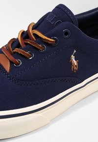 Námořnické plátěné tenisky s hnědými koženými akcenty, kontrastní bílou podrážkou a vyšitým logem. Ploché tkaničky a texturovaný povrch.