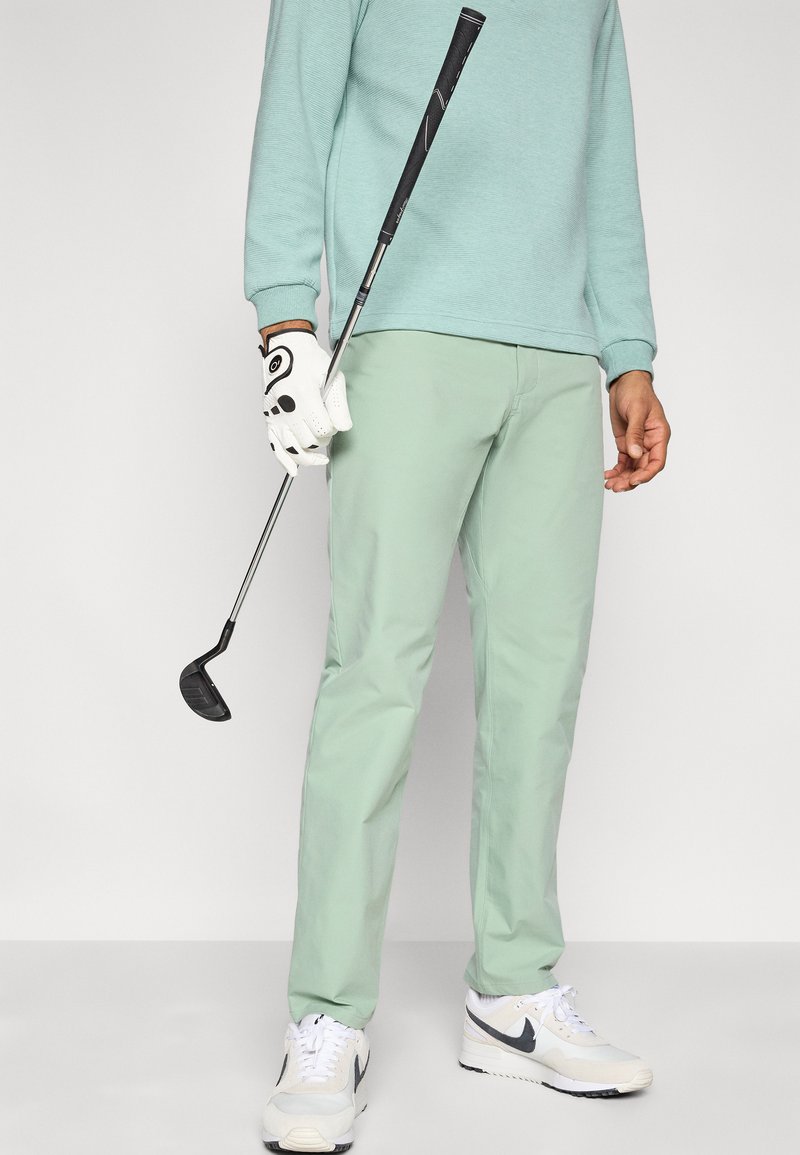 Lichtgroene rechte golfbroek, gecombineerd met een muntkleurige trui. Hand die een golfclub vasthoudt, met een witte handschoen met zwarte accenten.