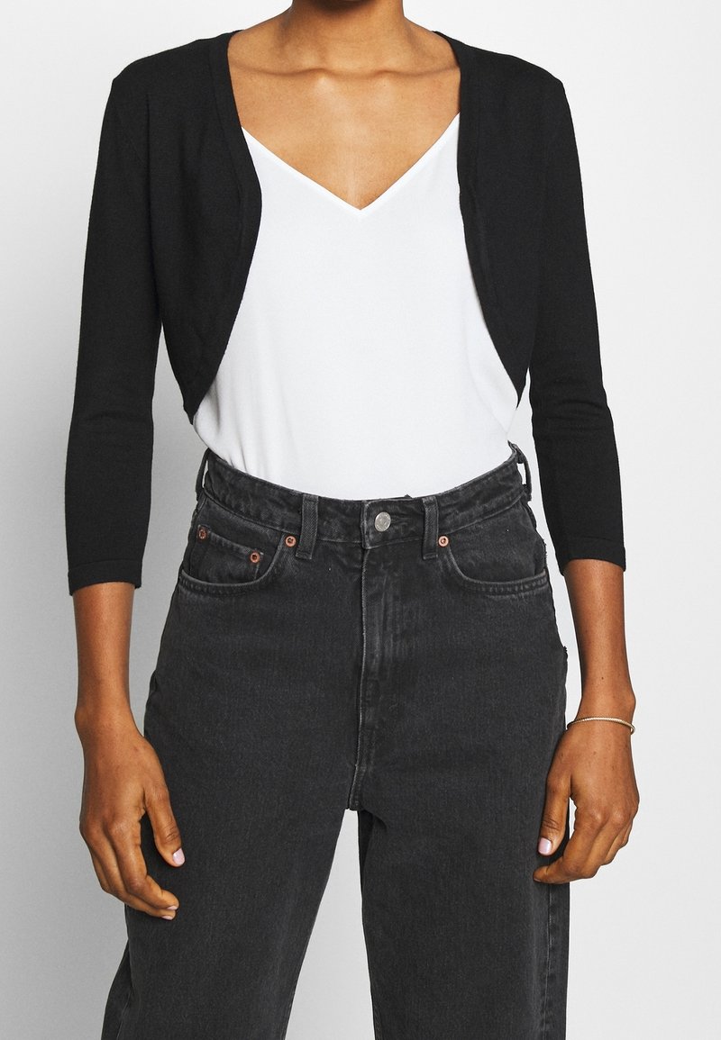 Mustan värinen lyhyt neuletakki pehmeästä kankaasta, jossa on kolme neljänneksen mittaiset hihat. Päällä on valkoinen V-kaula-aukkoinen paita, yhdistettynä korkeavyötäröisiin mustiin farkkuihin.