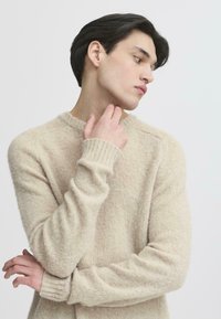 Suéter de punto beige con un acabado ligeramente texturizado, cuello redondo y puños acanalados. La modelo posa con la mano cerca de la barbilla frente a un fondo simple.