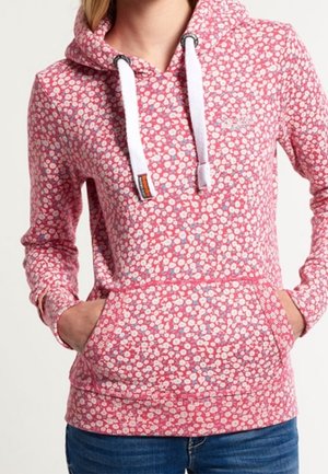 Frau trägt einen pinken Kapuzenpullover mit Blumenmuster und weißen Kordeln, Hände in der Vordertasche, kombiniert mit blauen Jeans.