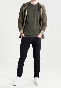 Olivgrüne Zip-Hoodie über einem dunkelgrünen gestreiften T-Shirt, kombiniert mit schwarzen Slim-Fit-Jeans und weißen Sneakern.