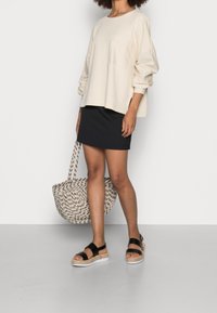 Sweatshirt beige oversize con maniche lunghe, dettaglio della tasca, abbinato a una gonna mini nera. Borsa a tracolla intrecciata e sandali neri con cinturini completano il look.