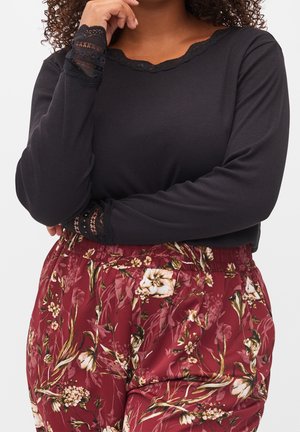 Vrouw draagt een zwart shirt met lange mouwen en kanten manchetten en een rode rok met bloemenpatroon, armen gekruist, hand bij het gezicht.