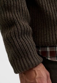 Primo piano di una mano che indossa una manica di maglione chunky a maglia marrone scuro sopra il polsino di una camicia a quadri rossa e bianca.