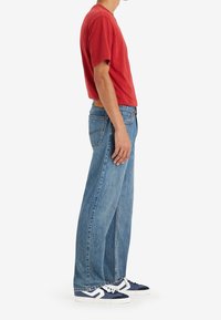 Personne portant un t-shirt rouge, un jean bleu large et des baskets bleu marine et blanches, debout de profil sur un fond blanc.
