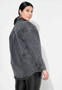 Chaqueta de mezclilla gris de gran tamaño con mangas largas, cuello y puños abotonados. Textura suave con un acabado ligeramente desgastado. Pantalones negros ajustados visibles.