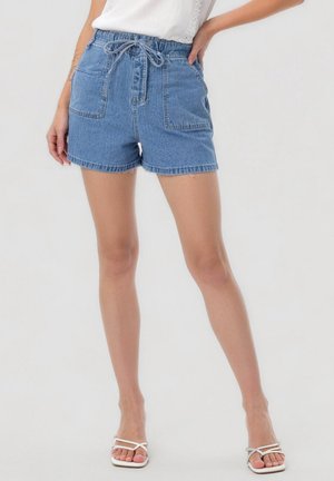 Femme portant un short en denim bleu avec une taille à cordon, deux poches plaquées devant, un haut blanc et des sandales blanches à lanières.
