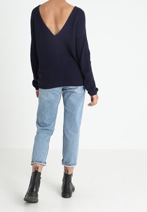 Personne portant un pull bleu marine à dos en V profond, un jean bleu clair retroussé et des bottines noires, debout dos à fond blanc.