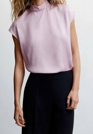 Femme portant un chemisier sans manches rose pâle à col montant rentré dans un pantalon noir taille haute, debout devant un fond uni.