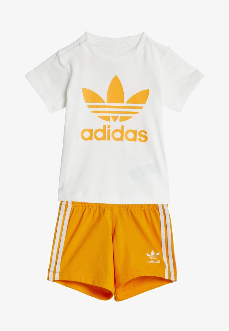 Camiseta blanca de algodón con un gran logo naranja de Adidas, combinada con pantalones cortos naranjas que cuentan con rayas blancas y un logo de Adidas en el dobladillo.