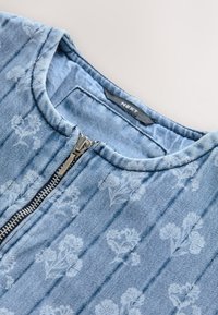 Denimtop i lyseblå med kviltet tekstur, med en forreste lynlås, rundet halsudskæring og et hvidt blomsterprint over det hele.