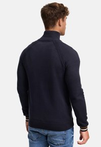 Tummansininen pullover korkealla kauluksella, teksturoitu neule, ribatut hihansuut ja helma, jotka korostuvat raitateemalla hihansuissa.