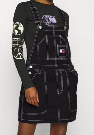 Persoon draagt een zwarte denim overall jurk met witte stiksels over een zwart topje met lange mouwen met peace-, globe- en Tommy Jeans-logo's.