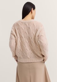 Gestrickter Pullover in einem sanften Rosa, mit einem Zopfmuster, überschnittenen Schultern und geripptem Saum. Kombiniert mit einem beigen Rock.