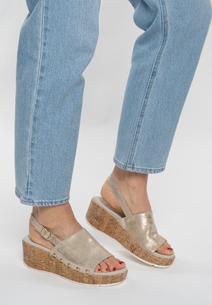Feet wearing light blue jeans and gold wedge sandals with cork soles and ankle straps on a white background.