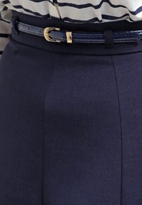 Kostymbyxor i marinblått med en slät yta, utrustade med ett smalt bälte med en guldfärgad metallspänne, som sitter vid midjan.